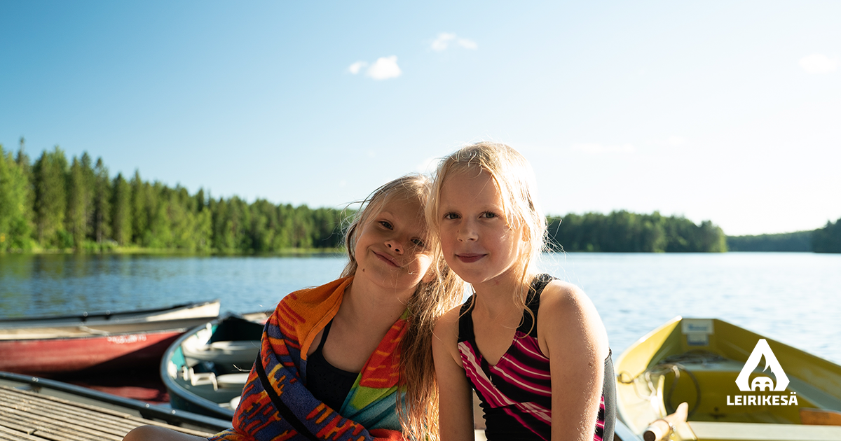 Kuvassa kaksi lasta istuvat laiturilla uintireissun jälkeen. Aurinko paistaa taustalta sinisellä taivaalla. 