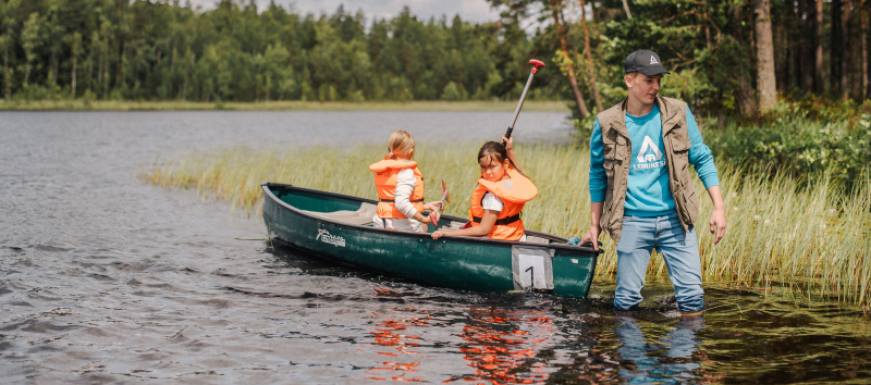 Työntekijä järven rannassa vetää leiriläisiä perässä kanootissa.