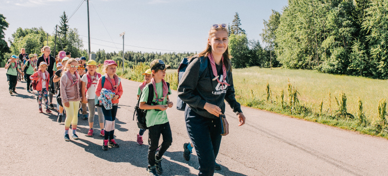 Työntekijä partiolaisten päiväleirillä kulkee jonossa leiriläisten kanssa.